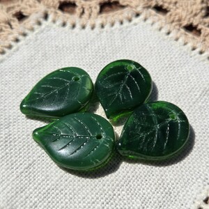 May include: Four translucent green glass leaf beads with etched vein details. Each bead has a small hole for stringing. The beads are arranged on a white fabric with a decorative lace border.