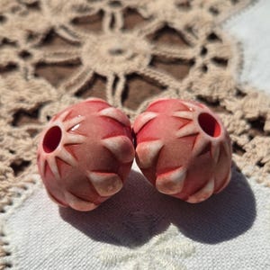 May include: Two pink ceramic beads with a white starburst design. The beads have a hole in the center for stringing.