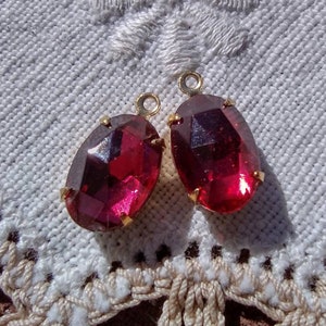 May include: Two oval-shaped red glass cabochons with gold-toned metal settings. The cabochons are set on a white crocheted surface.