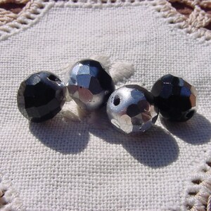 May include: Four round beads, two black and two silver, with a faceted design. The beads are on a white crocheted surface.