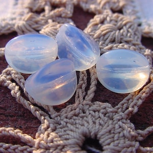 May include: Four iridescent, translucent, oval-shaped glass beads on a crocheted lace doily. The beads are a light blue color with a shimmery finish.