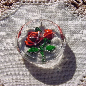 May include: A clear glass button with a red and green rose design. The button is round and has a raised design.