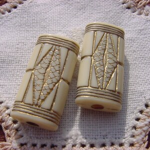 May include: Two white cylindrical beads with gold geometric designs. The beads have a smooth, polished finish.