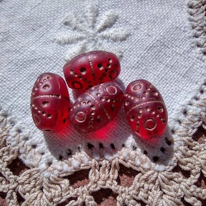 May include: Four red glass beads with gold accents and a patterned design. The beads are arranged in a pyramid shape on a white crocheted surface.