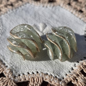May include: A pair of vintage clip-on earrings. Each earring features a layered, leaf-like design in pale green with gold-colored trim and white speckles. The earrings are displayed on a white, textured surface.