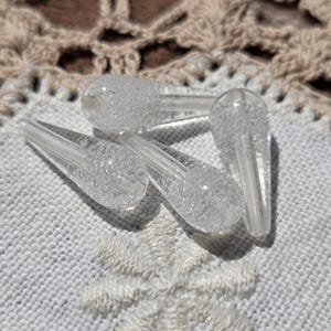 May include: Four clear glass teardrop-shaped beads with a textured surface. The beads are arranged in a cluster on a white crocheted surface.