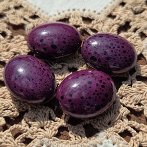 May include: Four speckled, purple decorative eggs. The eggs have a glossy finish and are covered in small, dark spots. They are arranged on a beige, crocheted doily with a floral pattern.