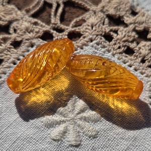 May include: Two amber-colored, leaf-shaped glass beads. The beads have a ribbed texture and are positioned on a white embroidered surface. The beads are casting shadows, and the background includes a crocheted doily.