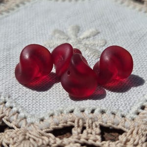 May include: Five translucent, heart-shaped, deep red glass beads. The beads are clustered together on a white, textured surface with a delicate, crocheted border. The beads have a smooth, polished appearance.