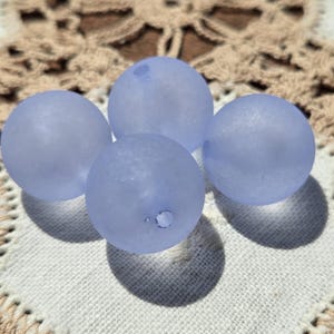 Può includere: Quattro perline di vetro azzurro smerigliato. Ogni perlina è rotonda con un piccolo foro. Le perline sono disposte su una superficie all'uncinetto bianca e beige, proiettando ombre morbide. Le perline hanno un aspetto leggermente strutturato.