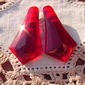 May include: Two red faceted glass teardrop-shaped pendants. The pendants are on a white crocheted doily.