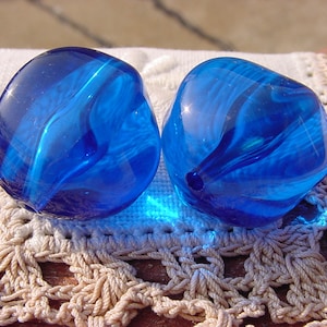 May include: Two blue glass beads with a swirled pattern. The beads are on a white crocheted surface.