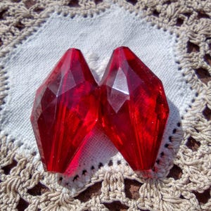 May include: Two red faceted glass beads, with a diamond shape, on a white crocheted doily.
