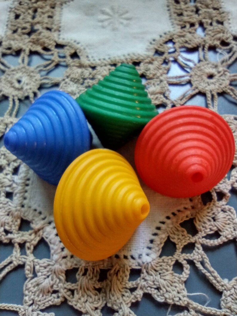May include: Four colorful spinning tops, one blue, one green, one yellow, and one red, are arranged on a white crocheted doily.