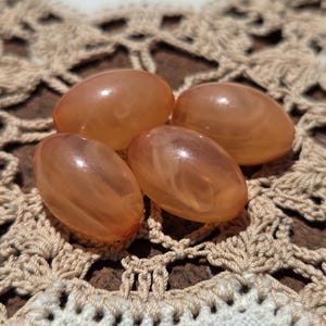 May include: Four translucent, oval-shaped beads in shades of peach and amber. The beads have a glossy finish and are arranged on a cream-colored, crocheted doily. The doily has a detailed, openwork pattern.