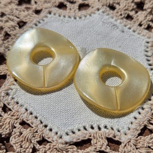 May include: Two large, round, pale yellow buttons with a hole in the center. The buttons have a smooth, shiny surface and a subtle shimmer.