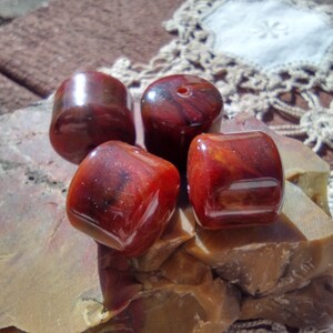 Pu&ograve; includere: Quattro perle rotonde marmoree di colore rosso e marrone con una finitura lucida. Le perle sono su una superficie ruvida, marrone e testurizzata.