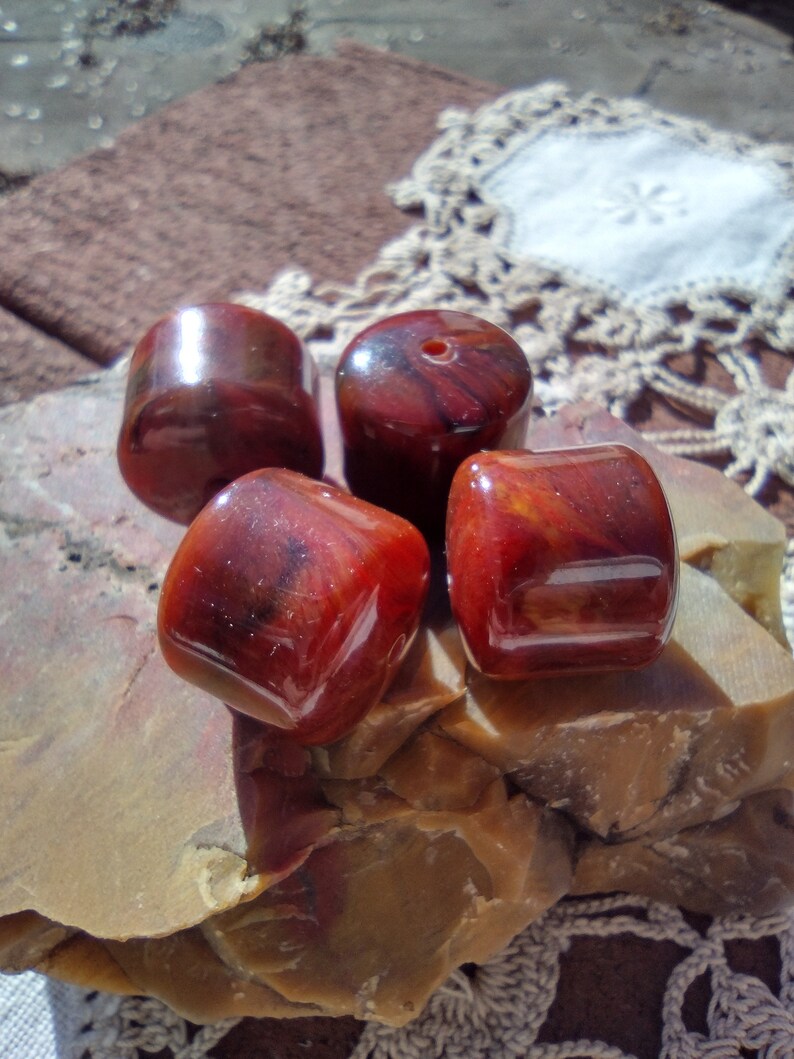 Pu&ograve; includere: Quattro perle rotonde marmoree di colore rosso e marrone con una finitura lucida. Le perle sono su una superficie ruvida, marrone e testurizzata.