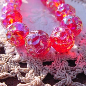 May include: A strand of iridescent pink glass beads with a textured surface. The beads are strung together on a white string and are lying on a crocheted doily.