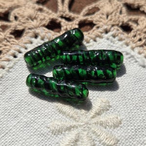 May include: Four green glass beads with black accents, arranged on a white embroidered surface. The beads have a cylindrical shape with a ribbed texture. The background includes a beige lace doily.