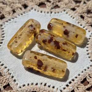 May include: Four rectangular, amber-colored beads with brown inclusions are arranged on a white crocheted doily. The beads have a translucent quality, and the doily has a delicate, openwork design.