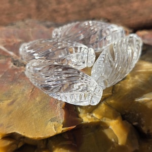 May include: Four clear, glass, oval-shaped beads with a textured, ridged design. The beads are resting on a multi-colored, rough-textured stone surface. The beads are transparent, allowing light to pass through.