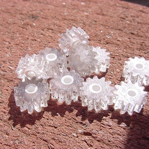 May include: A group of small, clear plastic gears with white centers. The gears are arranged in a pile on a red brick surface.