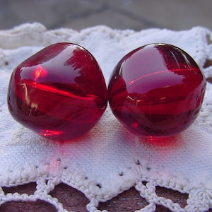 May include: Two large, round, red glass beads. The beads are smooth and shiny.