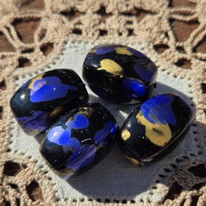 May include: Four black glass beads with blue and gold accents. The beads have an oblong shape and are arranged on a cream-colored, crocheted surface. The beads have a glossy finish and are likely used for jewelry making.