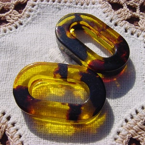 May include: Two oval-shaped, amber-colored plastic toggles with black accents. The toggles are on a white crocheted surface.