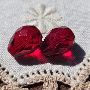 May include: Two faceted, teardrop-shaped, deep red glass beads. The beads are positioned on a white, textured surface with a floral design and a cream-colored, crocheted border. The beads cast a shadow.