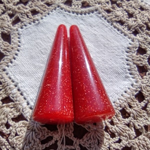 Può includere: Due oggetti rossi a forma di cono con macchie simili a glitter. Sono appoggiati su un tessuto bianco con un centrino beige all'uncinetto. Si tratta probabilmente di oggetti decorativi o funzionali.