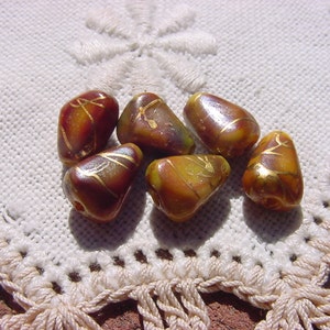 May include: Six brown, teardrop-shaped beads with gold-colored accents. The beads have a glossy finish and are arranged on a white, crocheted surface. The beads have a unique, organic shape and are likely used for jewelry making.