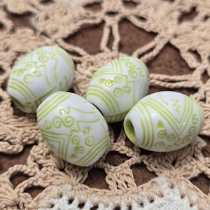 May include: Four white, oval-shaped beads with a light green, decorative pattern. The beads are arranged on a beige, crocheted surface. The beads have a hole running through the center.