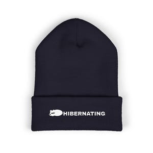 Könnte beinhalten: Eine dunkelblaue Mütze mit Umschlag, die eine weiße Stickerei der Silhouette eines schlafenden Bären zeigt, gefolgt von dem Text HIBERNATING in einer serifenlosen Schrift, zentriert auf dem vorderen Umschlag des Strickstoffs.