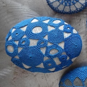May include: A close-up of a round, gray stone covered in a blue crocheted design. The design features circles, triangles, and other geometric shapes.