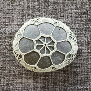 Puede incluir: Una piedra gris está adornada con un delicado tapete de ganchillo verde claro. El tapete presenta un diseño floral con una estrella central y patrones circulares, creando una pieza decorativa única. El fondo es una superficie texturizada marrón.