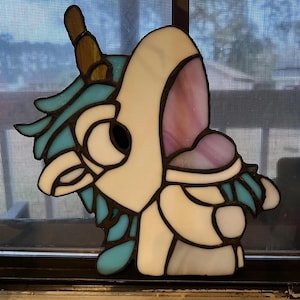 Peut inclure: Décoration de fenêtre en vitrail représentant une licorne. La licorne a un corps blanc, une crinière et une queue turquoise, et une corne dorée. L'objet est posé devant une fenêtre.