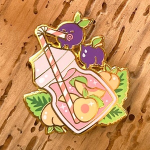 Peut inclure: Une épingle en émail rose et or représentant une bouteille en verre avec une paille, remplie de tranches de pêche et d'une myrtille. La bouteille est entourée de feuilles vertes.