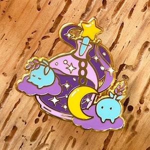 Peut inclure: Une épingle en émail violette et dorée représentant une bouteille avec une étoile au sommet, une lune croissante et deux petites créatures bleues aux yeux blancs. La bouteille est remplie d'étoiles et une chaîne pend du sommet.