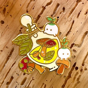 Peut inclure: Une épingle en émail dorée représentant une bouteille en verre remplie de champignons, de feuilles et d'étoiles brillantes. La bouteille est surmontée d'un petit personnage blanc en forme de champignon avec une pousse verte. Il y a un deuxième petit personnage en forme de champignon sur le côté droit de la bouteille.