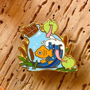 Peut inclure: Épinglette en émail doré représentant un poisson de dessin animé, deux grenouilles et une barre de chocolat dans un design de vague bleu et blanc.