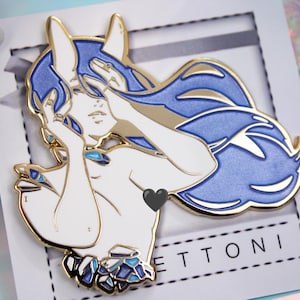 May include: A gold-colored enamel pin featuring a stylized figure with blue hair and accents. The figure is depicted with their hands near their face, with a heart shape on the chest. The pin is on a white card with the word "ETTONI" printed on it.