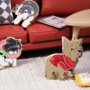 May include: Enamel pins featuring dog illustrations. One pin depicts a dog wearing a red sweater, another shows a dog with a pink collar, and a third shows a dog lying down. A bag of potato chips is also visible.