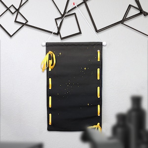 May include: Black wall hanging with gold ribbon accents. The rectangular banner has a dotted pattern and gold star details. The banner is suspended from a white rod with a white cord.