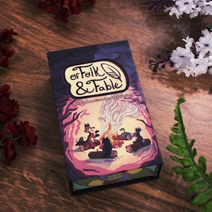 May include: A rectangular box for a collaborative card project, "of Folk & Fable", featuring a colorful illustration of a campfire scene with animal characters. The box is set on a wooden surface with floral accents.