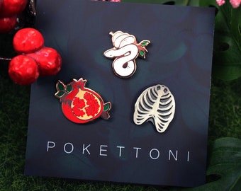 Puede incluir: Tres pines de esmalte sobre un fondo oscuro. Los pines presentan una serpiente blanca con sombrero, una granada roja y una caja torácica. La palabra "POKETTONI" está impresa en la parte inferior de la tarjeta.