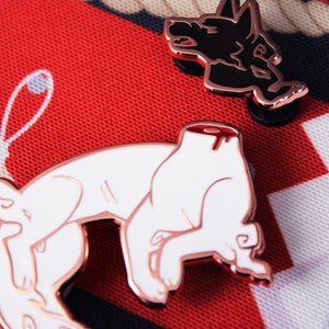 May include: Two enamel pins depicting a dog. One pin is black and shows a dog's head with its mouth open. The other pin is white and shows a dog lying down with its head cut off and a red drip of blood.