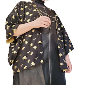 May include: Black kimono-style jacket with gold eye and eyelash pattern. The jacket has wide sleeves and a gold lining. A gold chain necklace and a black leather dress with a zipper are also visible.