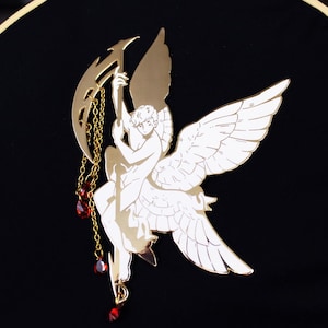 Pode incluir: Um pingente de metal dourado com um anjo estilizado com as asas abertas, segurando uma lua crescente com contas de cristal vermelho penduradas.